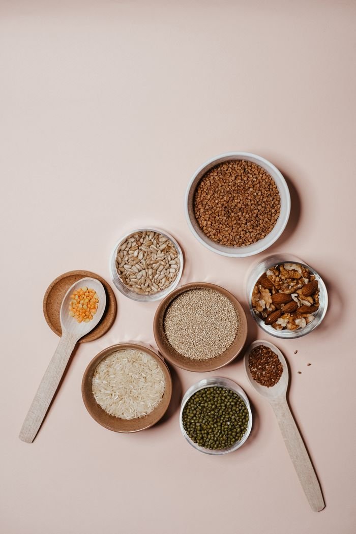 Flat lay of various grains and nuts in bowls and wooden spoons on neutral background.